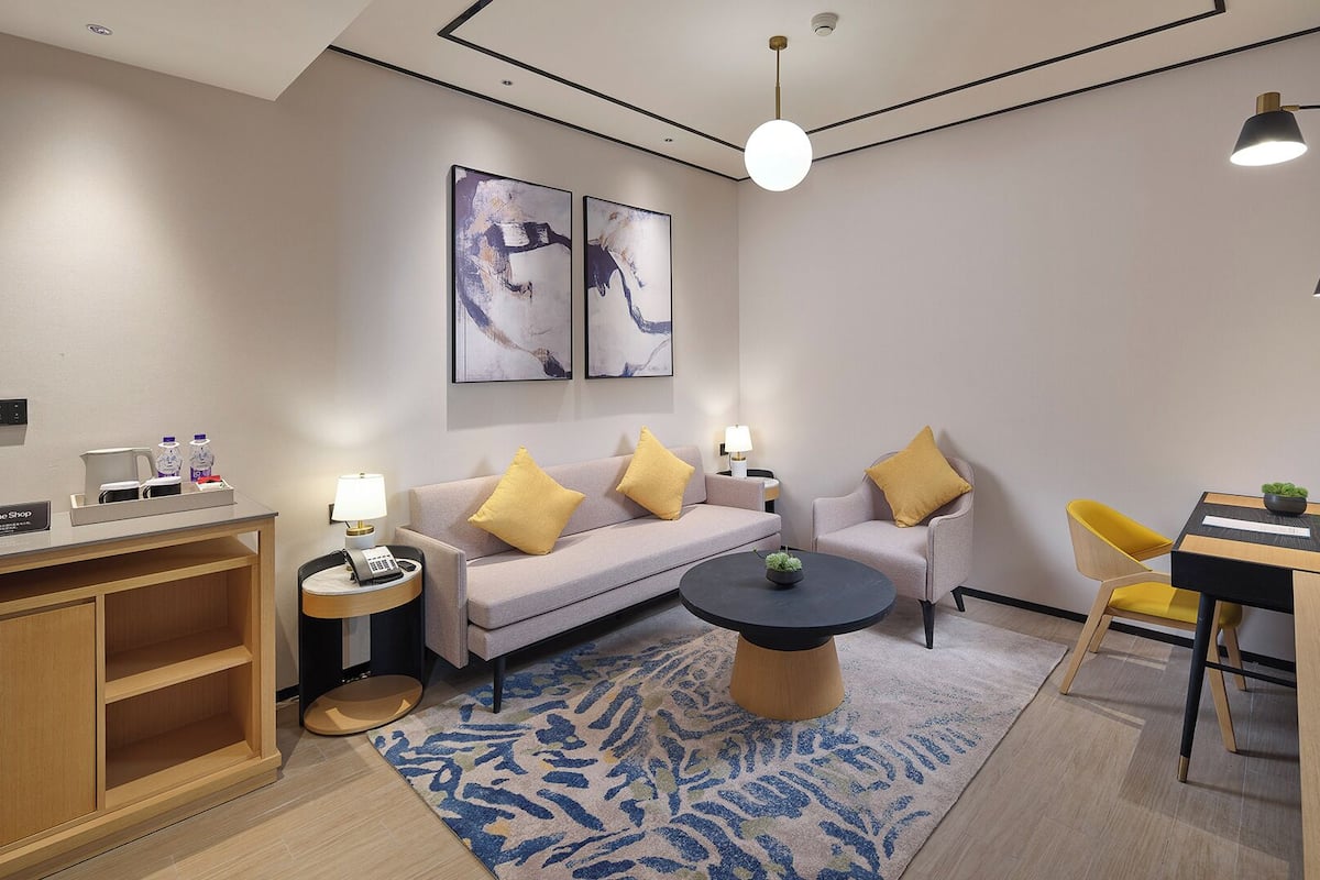 A modern hotel room with a sofa, a coffee table, a desk, and a television. There are two paintings on the wall, a small plant on the coffee table, and a rug on the floor.
