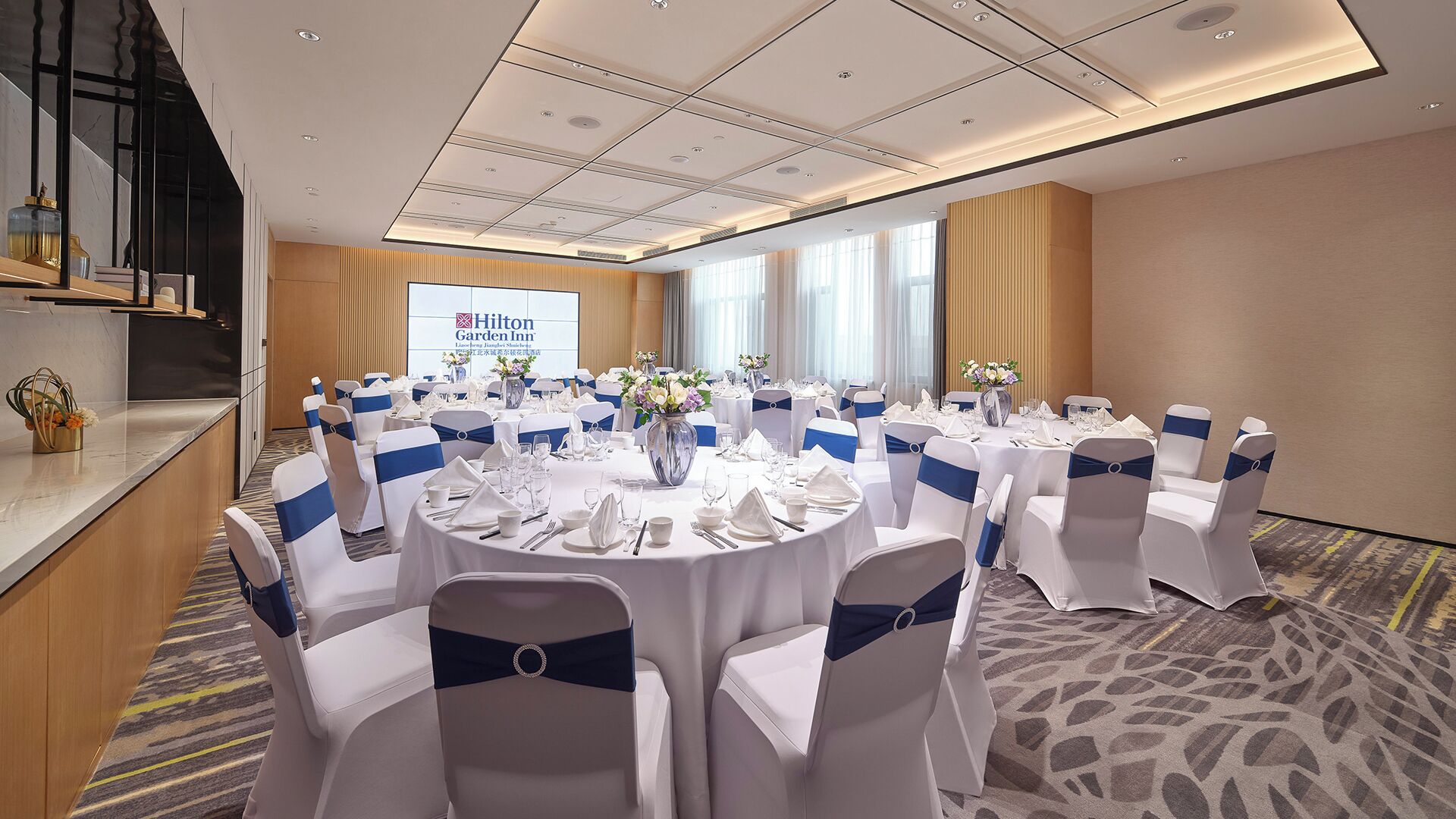 A conference room at Hilton Garden Inn with round tables, white chairs with blue sashes, and a projection screen displaying the hotel's name.