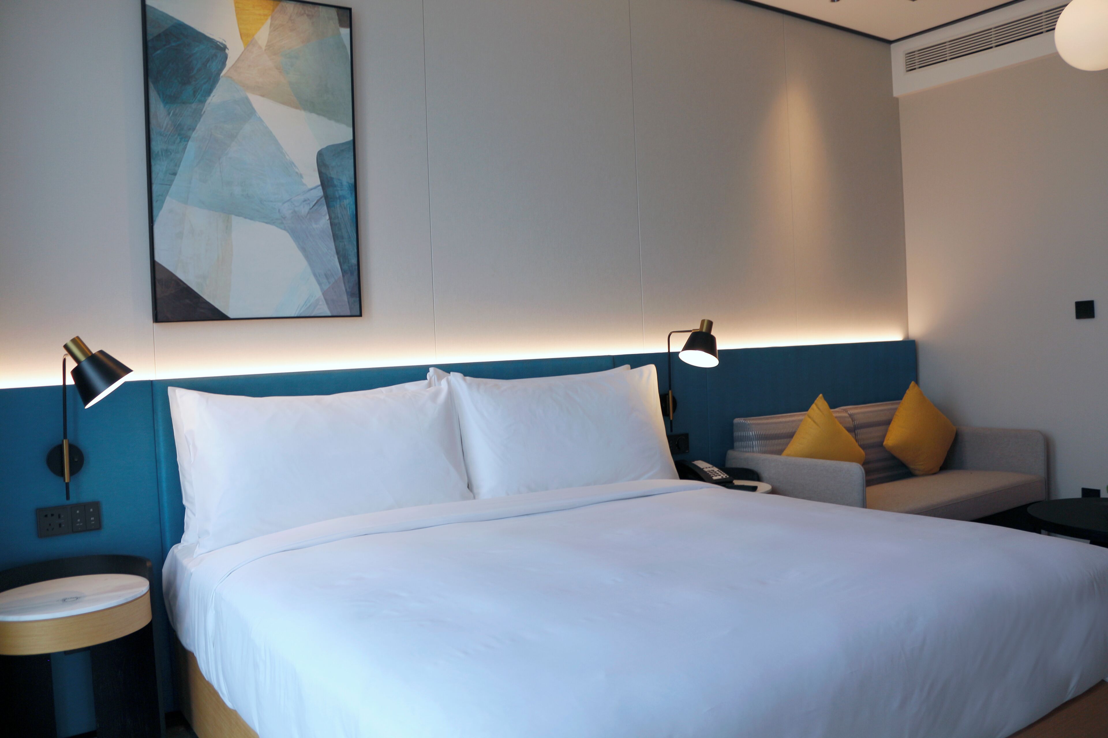 A hotel room with a bed, a nightstand, a lamp, a sofa with cushions, and a wall art piece.