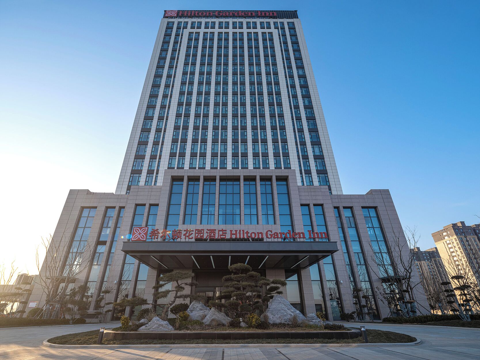 Un grand immeuble moderne, avec un ciel dégagé. L’immeuble porte une enseigne sur laquelle on peut lire « Hilton Garden Inn ».