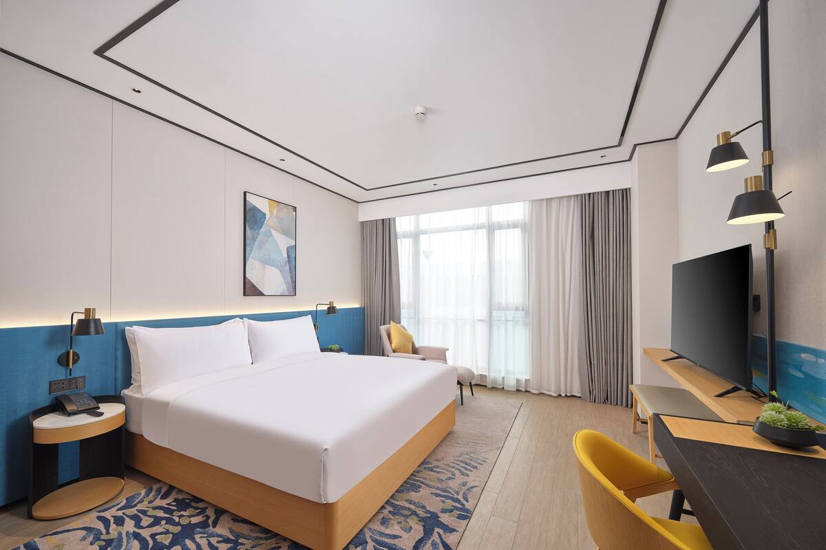 A modern hotel room with a large bed, bedside tables with lamps, a desk with a TV, and a view of the outdoors through large windows.