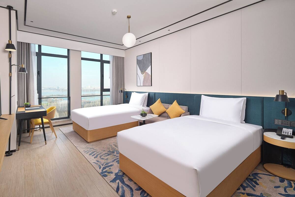 A hotel room with two beds, a desk, a TV, and a view of the city.