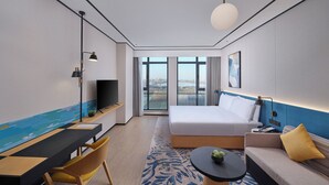 A modern hotel room with a large bed, a desk with a TV, a sofa, and a view of the city through large windows. - Hilton Garden Inn Liaocheng Jiangbei Shuicheng (Liaocheng)
