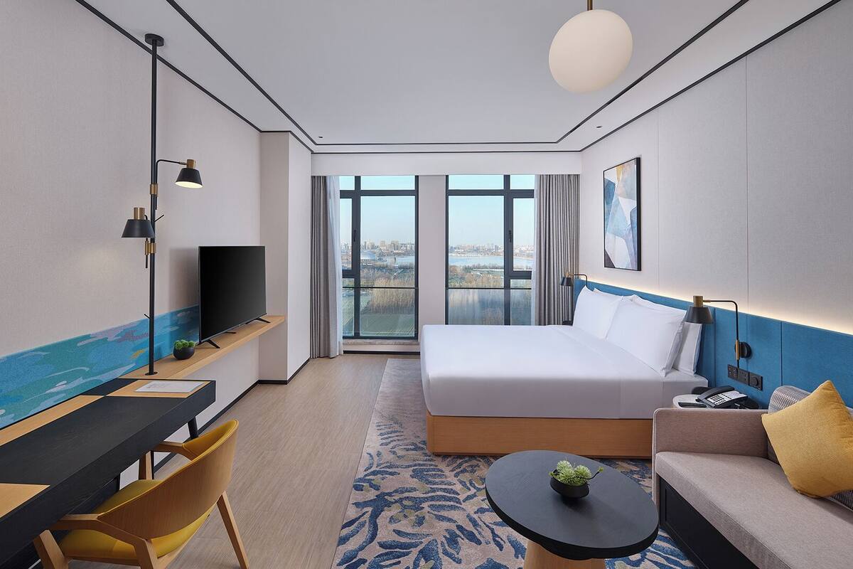 A modern hotel room with a large bed, a desk with a TV, a sofa, and a view of the city through large windows.