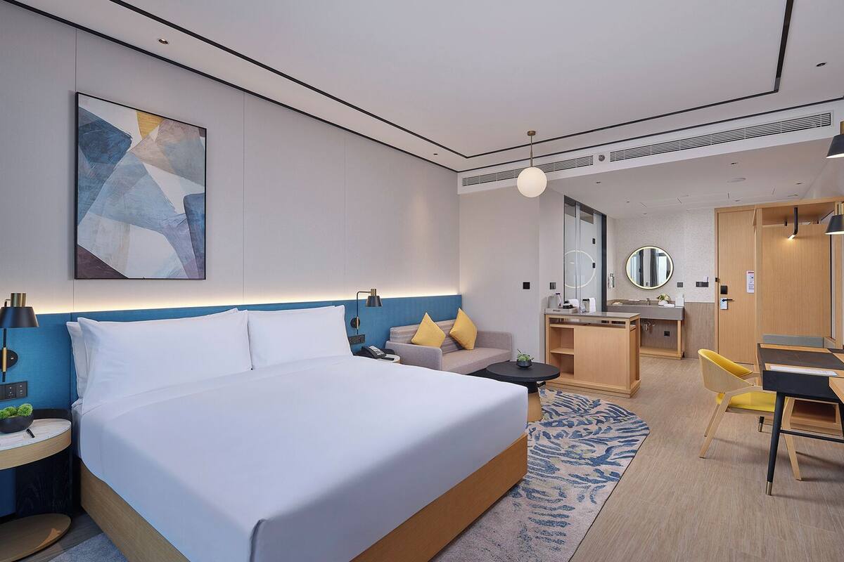 A modern hotel room with a large bed, a desk, a chair, a small sofa, a TV, and a bathroom with a mirror and sink.