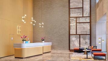 Un moderno vestíbulo de hotel con un mostrador de recepción de madera, una zona de estar con sofá y sillones, y un gran ventanal con diseños intrincados.