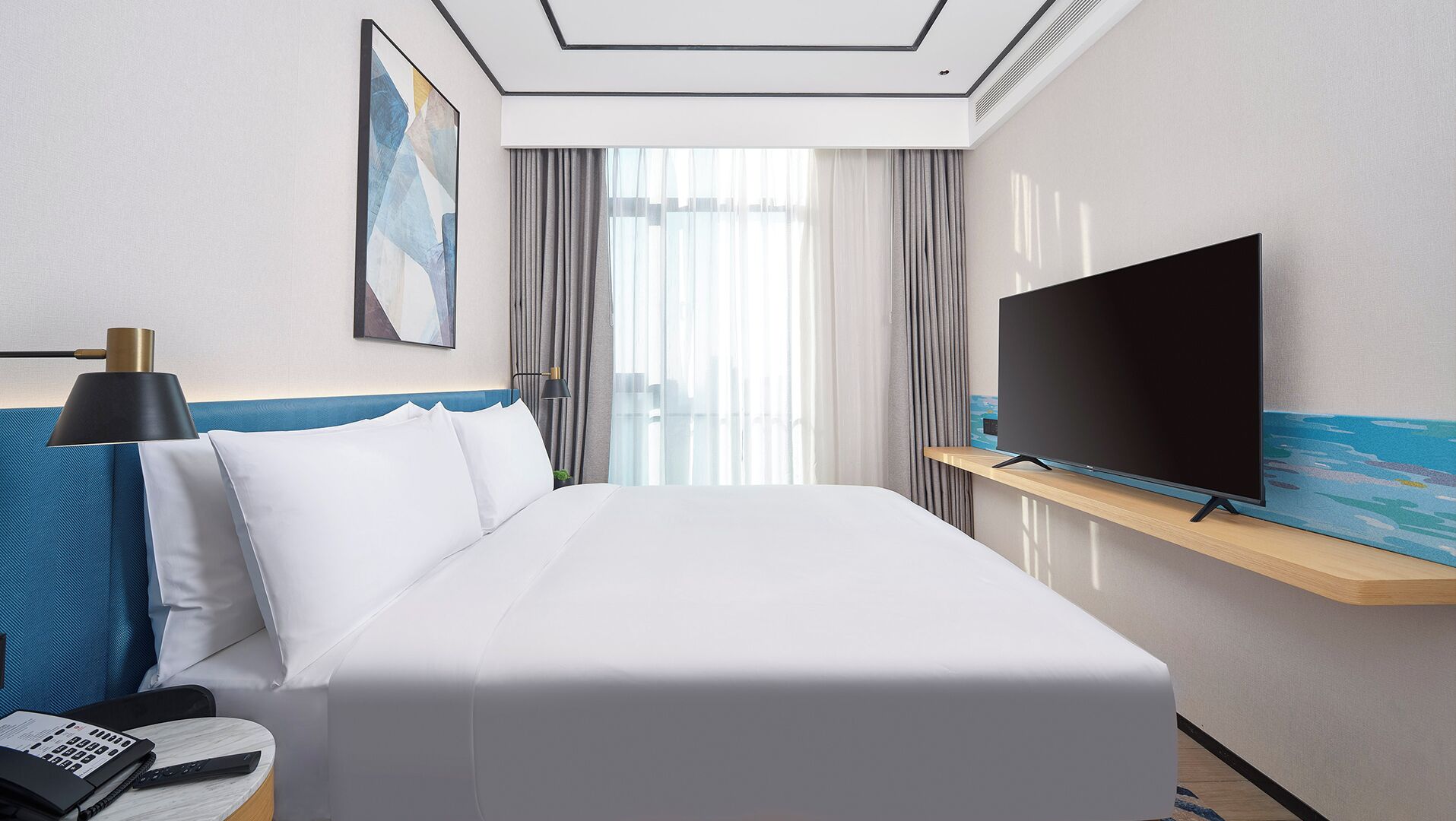 A hotel room with a bed, a TV on a wooden shelf, a desk with a lamp, and a painting on the wall.