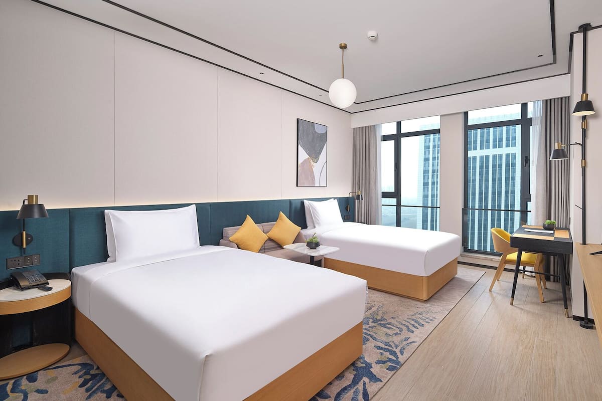A hotel room with two beds, a desk, a chair, a TV, and a large window with a city view.