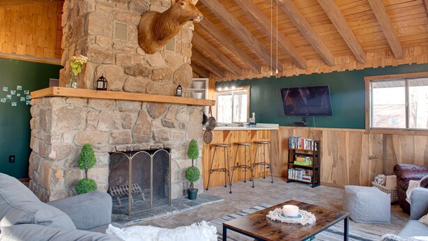 Ein rustikales Interieur mit Kamin aus Stein, Holzbalken, einem aufgehängten Hirschgeweih, einer Bar und einem Fernseher.