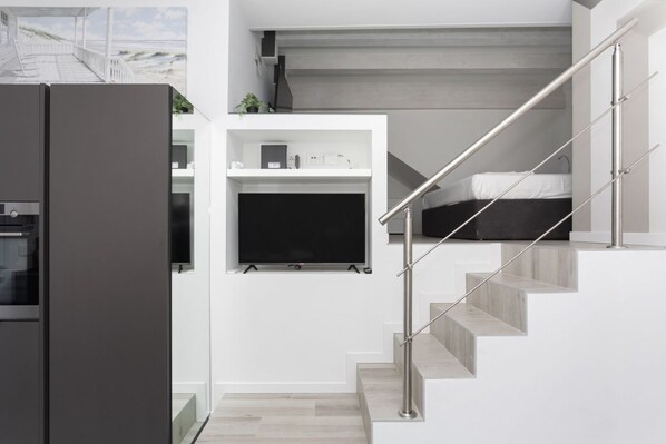 A modern kitchen with a built-in oven, a white wall-mounted TV, and a staircase with a metal railing.