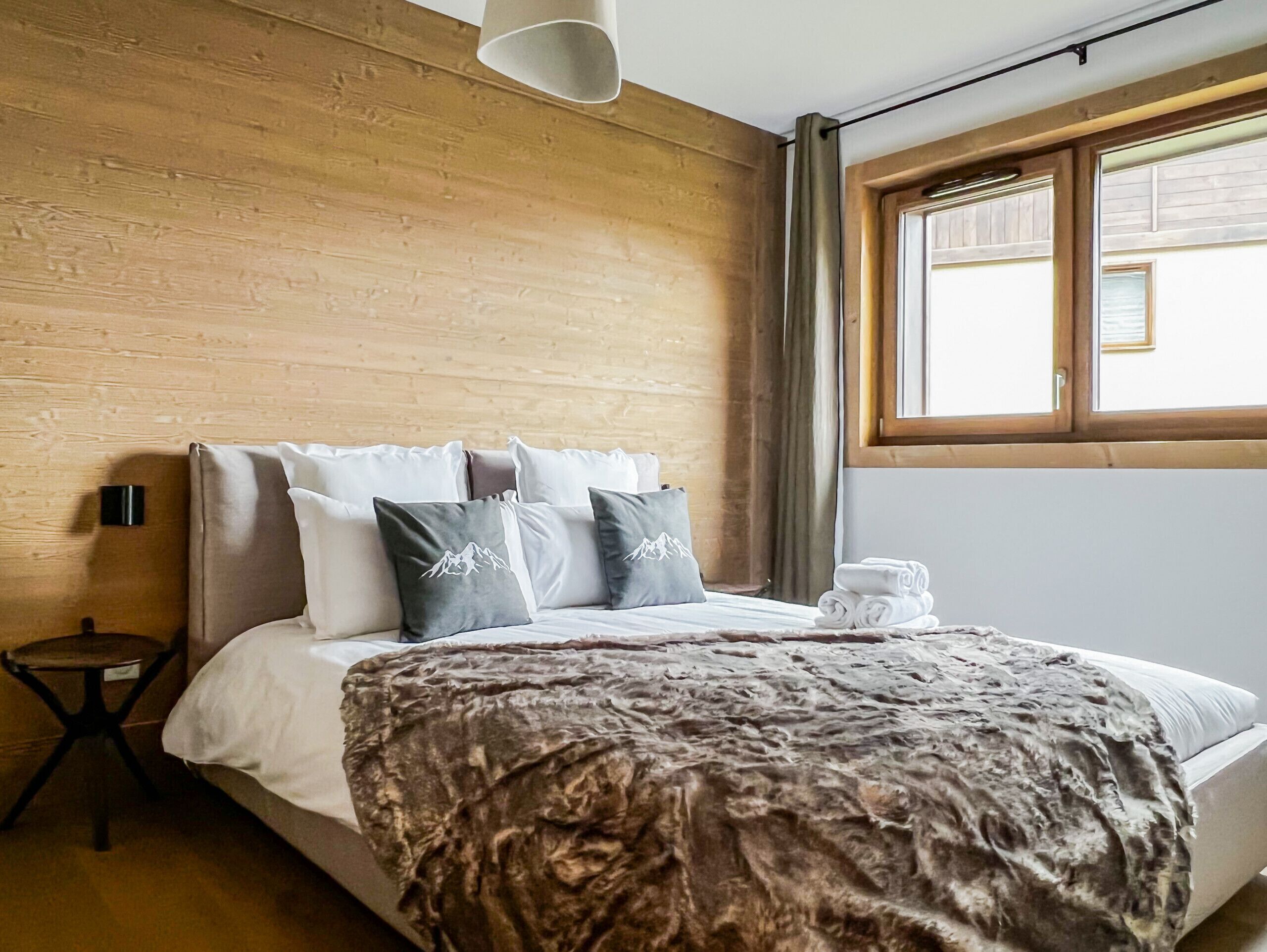 Habitación de hotel con pared de madera, cama con almohada de motivos de montaña, una mesita redonda y un ventanal con cortinas.