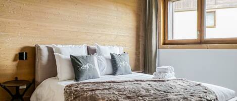 Habitación de hotel con pared de madera, cama con almohada de motivos de montaña, una mesita redonda y un ventanal con cortinas.
