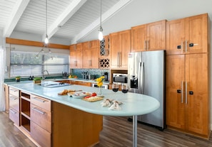 A modern kitchen with wooden cabinets, a stainless steel refrigerator, and a large island with food items and wine glasses.