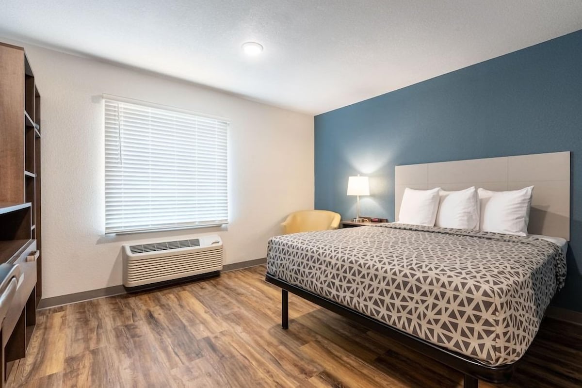 A hotel room with a bed, a chair, a desk, a lamp, and a window with blinds.