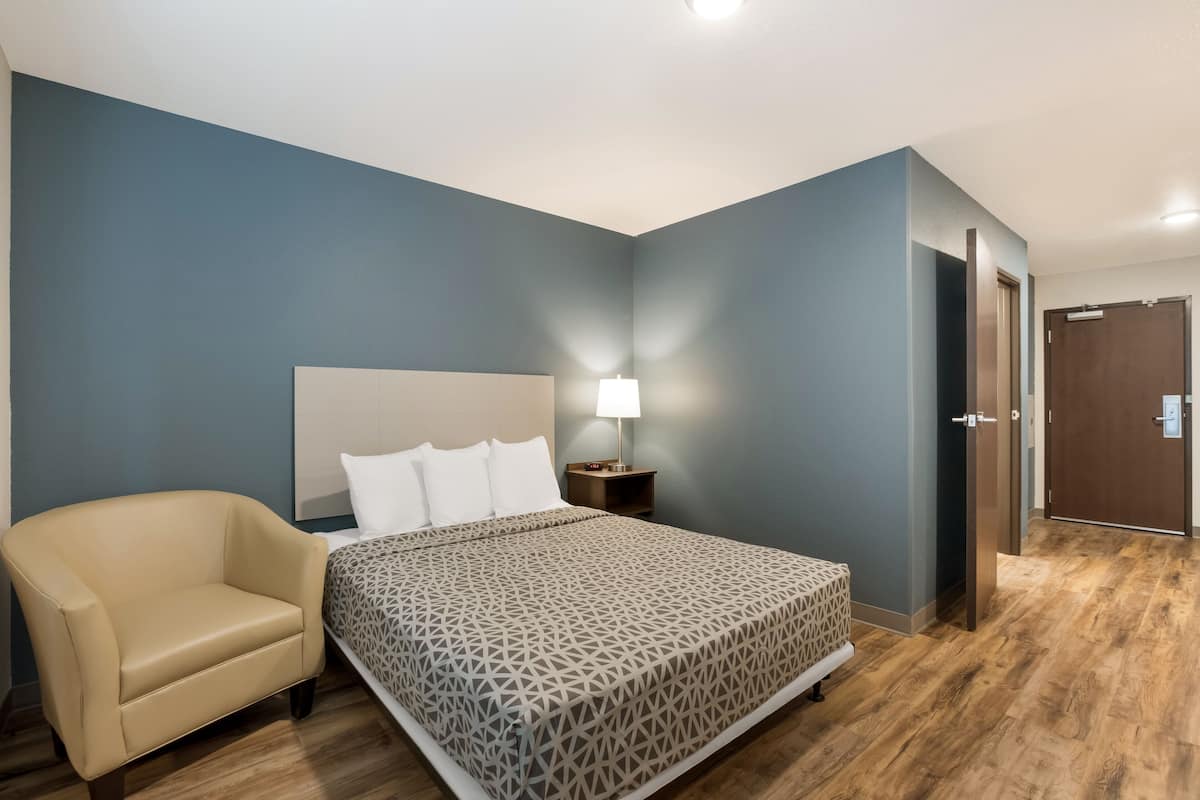 A hotel room with a bed, a chair, a nightstand with a lamp, and a door.