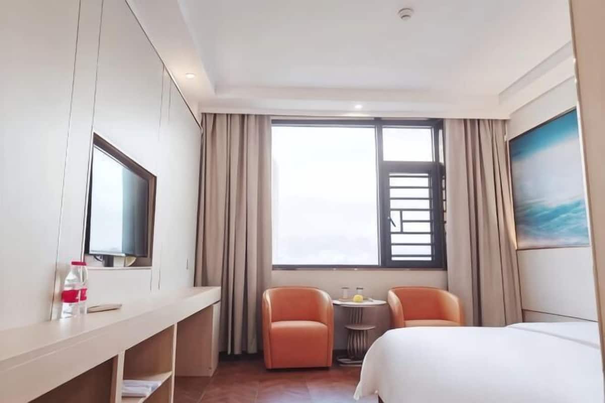 Quarto de hotel com cama, televisão, secretária com cadeira, pequena mesa com duas cadeiras e uma janela grande com cortinas.