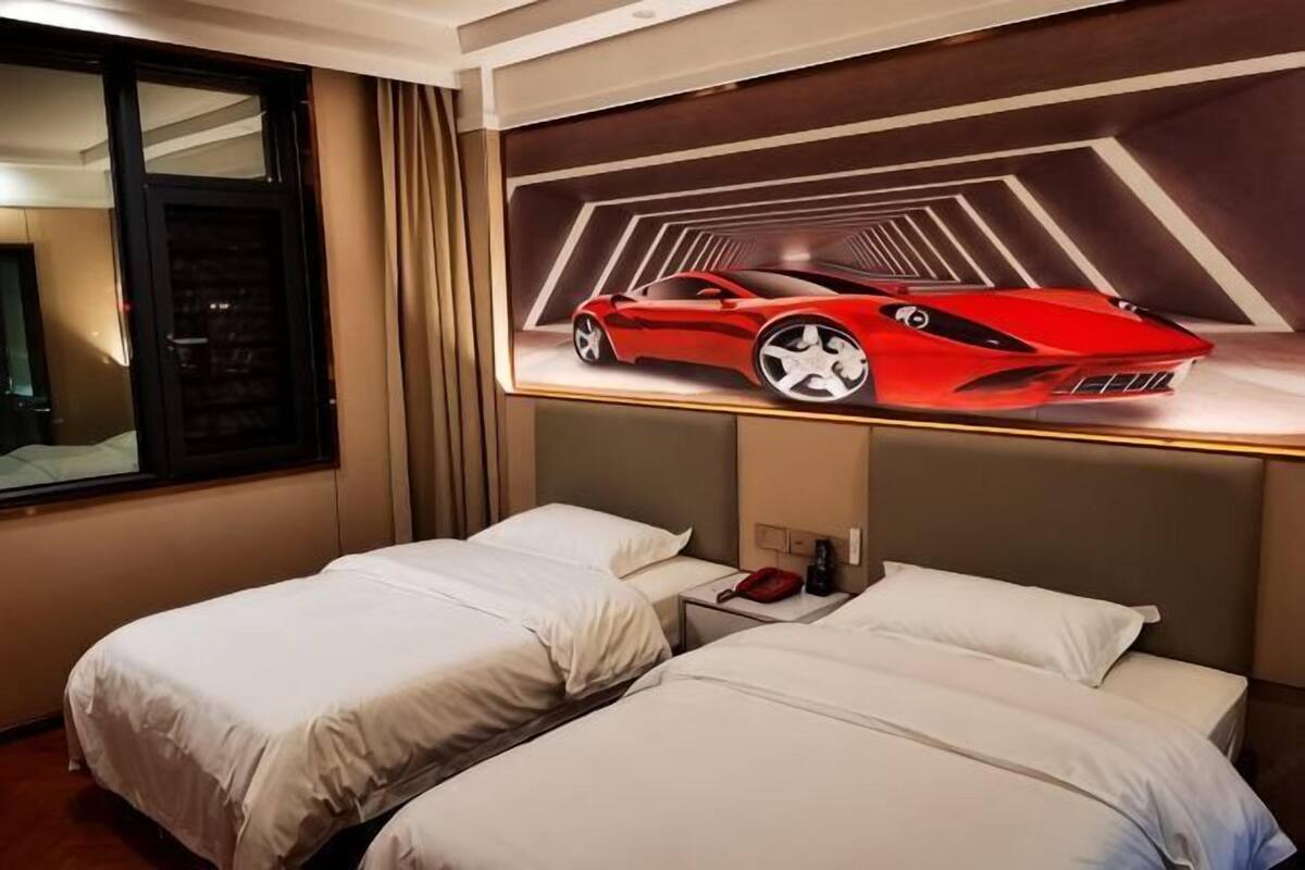 Quarto de hotel com duas camas, uma janela grande e uma televisão na parede mostrando um carro esportivo vermelho.
