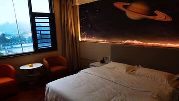 A hotel room with a large window, a bed, a small round table, two armchairs, and a wall mural of a planet with rings.