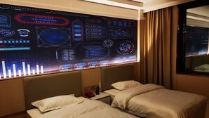 Habitación de hotel con dos camas, una mesita de noche y una pantalla digital grande en la pared.