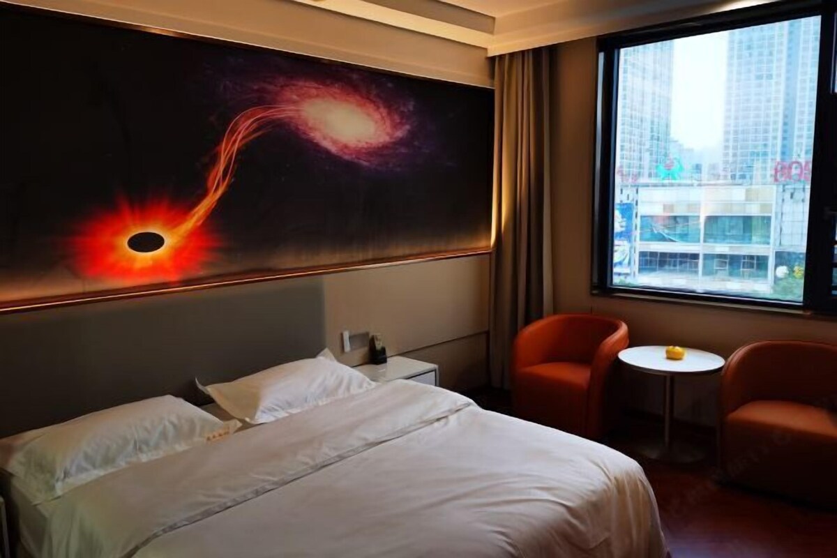 A hotel room with a large bed, two armchairs, a small table, and a painting of a galaxy.