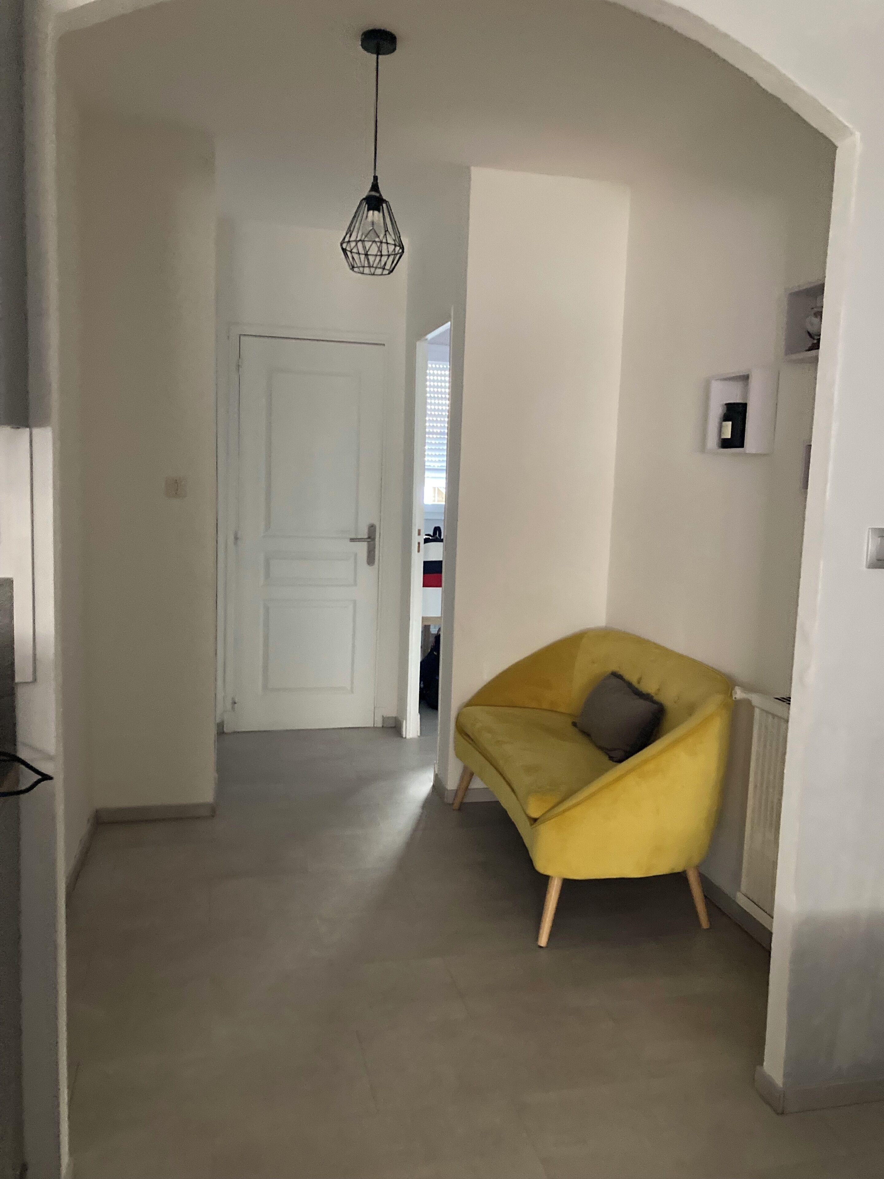 Un couloir avec une porte blanche, un fauteuil jaune avec un coussin et une suspension.