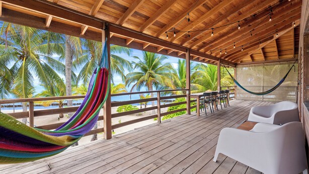 Una terraza de madera con una hamaca, sillas reclinables blancas y vistas a palmeras y al mar.