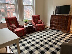 A room with a checkered floor, two armchairs, a wooden dresser, a television, and a window with a view of a brick building.