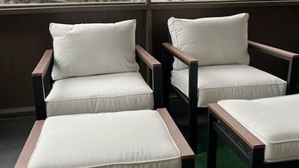 A balcony with three wooden chairs and cushions, a green artificial turf floor, and a view of trees and buildings through large windows.