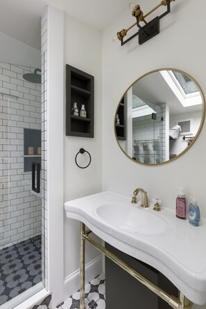 A bathroom with a white sink, gold fixtures, a round mirror, and a shower with patterned tiles. - Charming Yellow Cottage in Dilworth! (Charlotte)