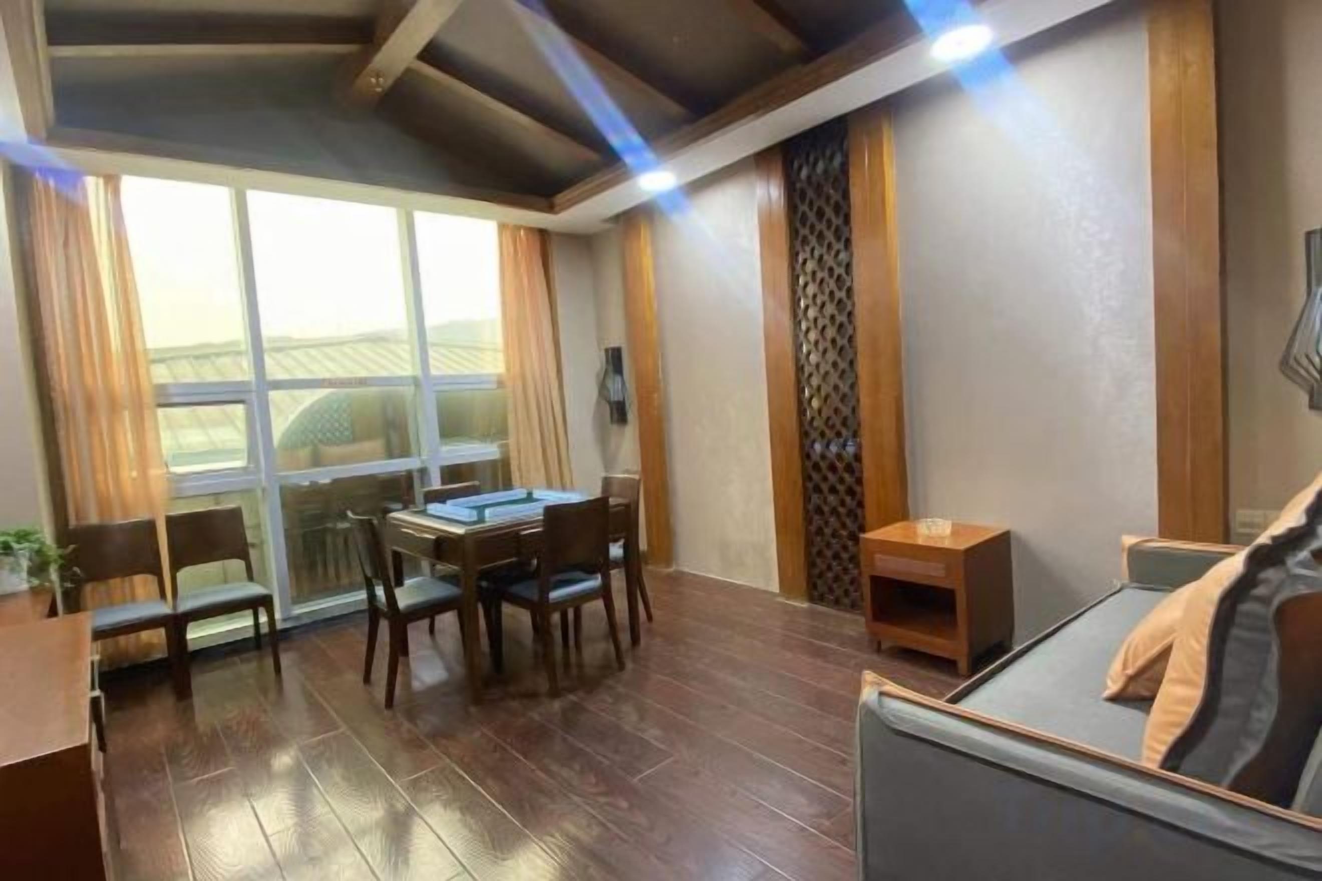 A room with a dining table, chairs, a sofa, a small wooden side table, and a large window with curtains.