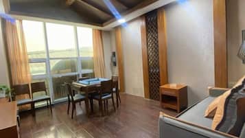 A room with a dining table, chairs, a sofa, a small wooden side table, and a large window with curtains.
