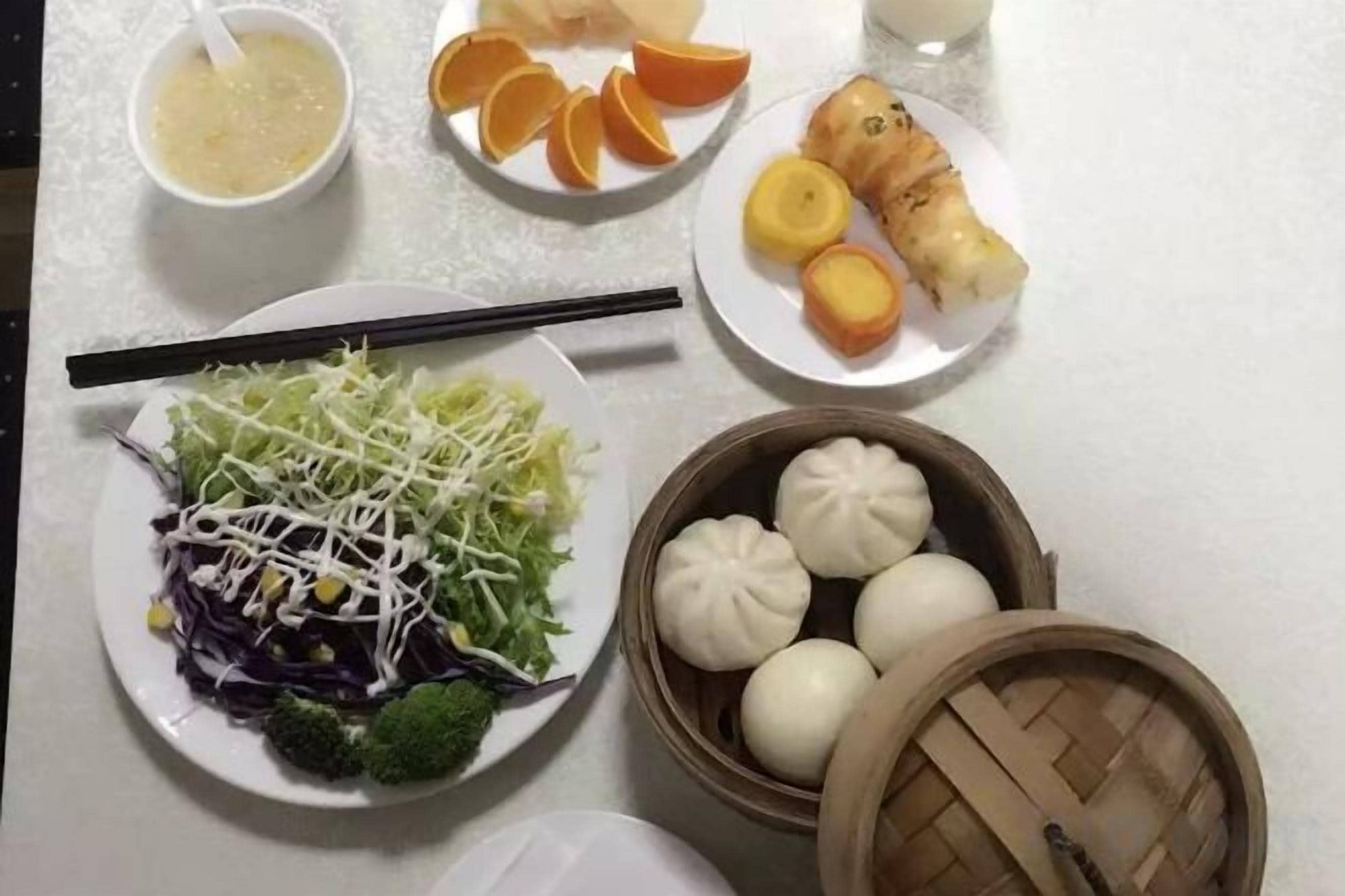 Una mesa con diversos alimentos, incluyendo un tazón de sopa, mandarinas en rodajas, un plato con una fruta amarilla, un plato con una ensalada de vegetales verdes y un plato con dumplings blancos.