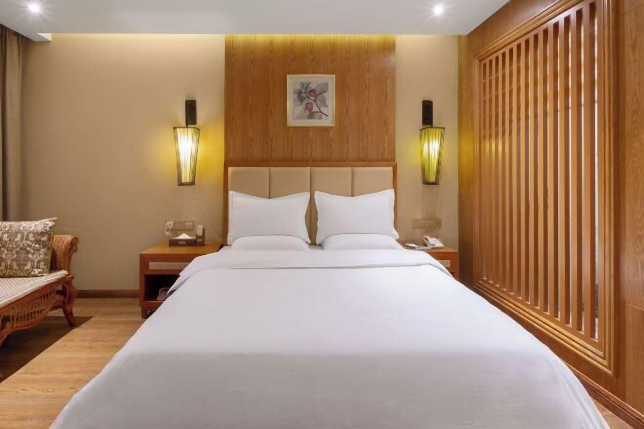 A hotel room with a large bed, wooden headboard, two bedside tables with lamps, a wooden wardrobe, and a wall-mounted artwork.