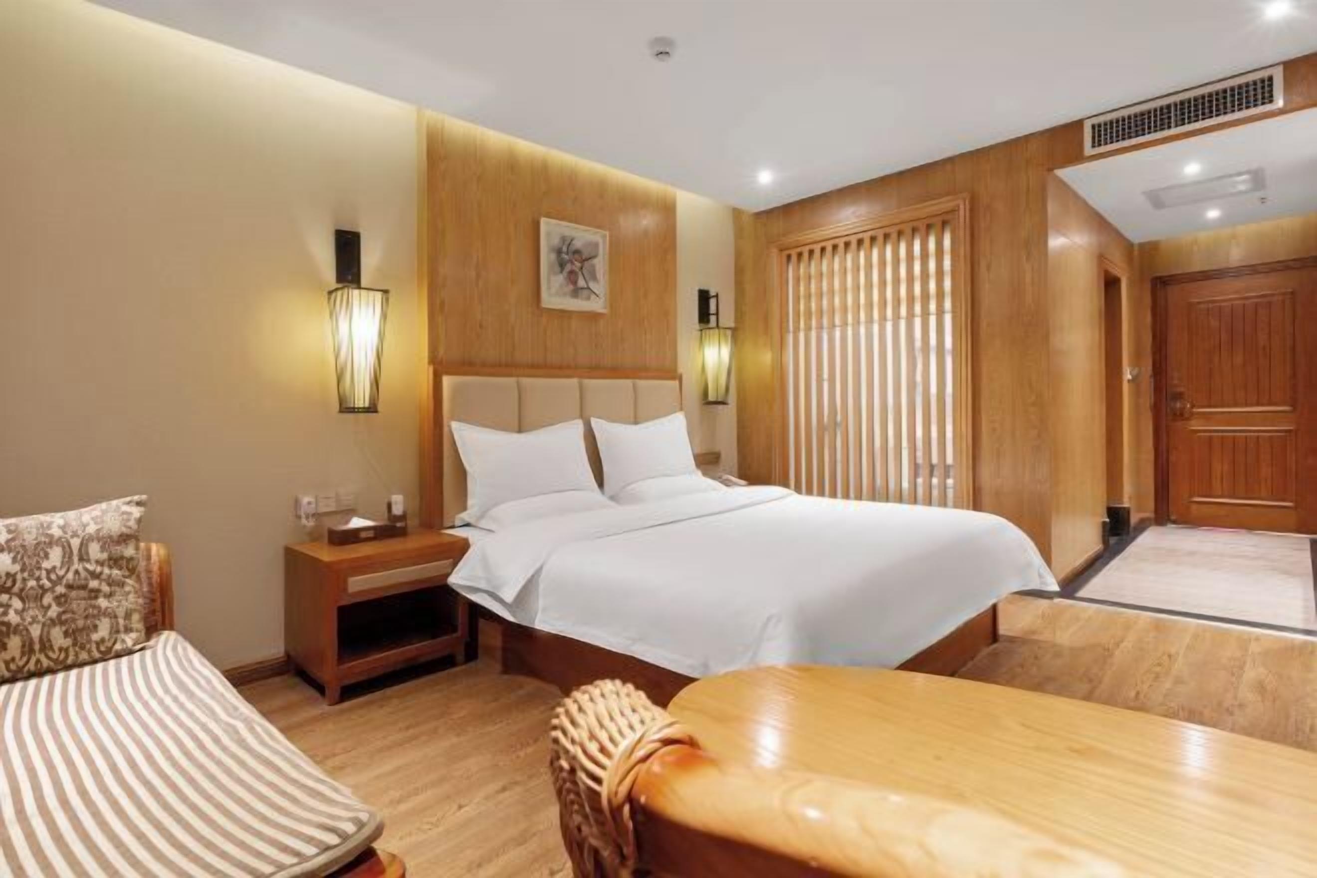 A hotel room with a large bed, a wooden nightstand, a bedside table, a chair, and a dining table.
