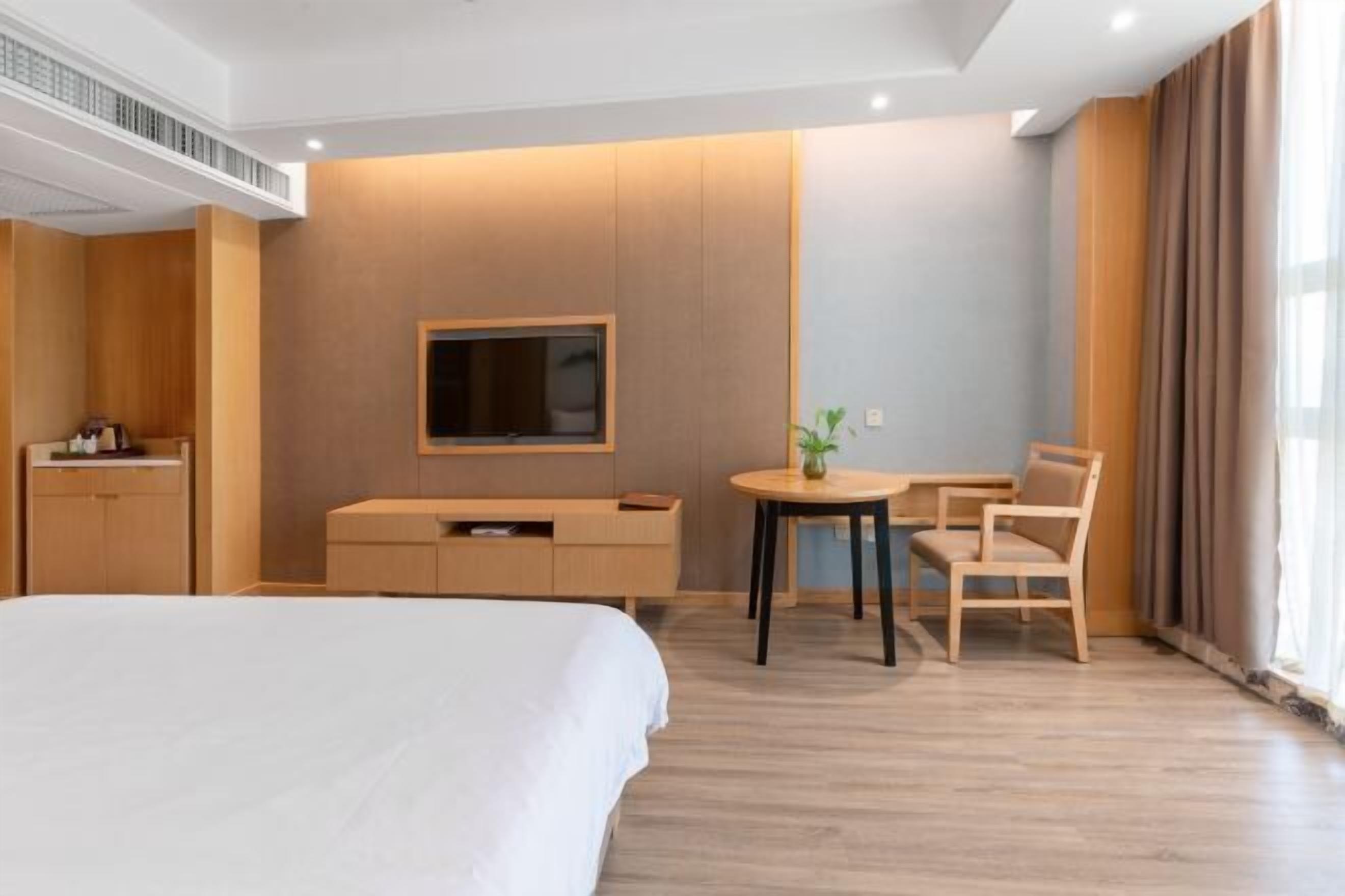 A modern hotel room with a large bed, a TV mounted on the wall, a small table with chairs, and a desk with a chair.