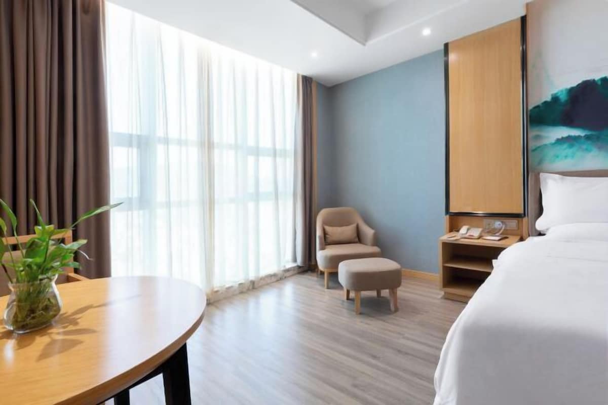 A hotel room with a bed, a chair, a small table with a plant, a nightstand, and a painting on the wall.