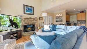 A living room with a stone fireplace, a large flat-screen TV, a blue sectional sofa, and a dining area in the background. - Fairway to Seven - King Bed, Big-screen TVs & Golf Course Views (Jasper)