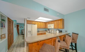 A kitchen with wooden cabinets, a microwave, a refrigerator, and a bar area with wicker chairs.