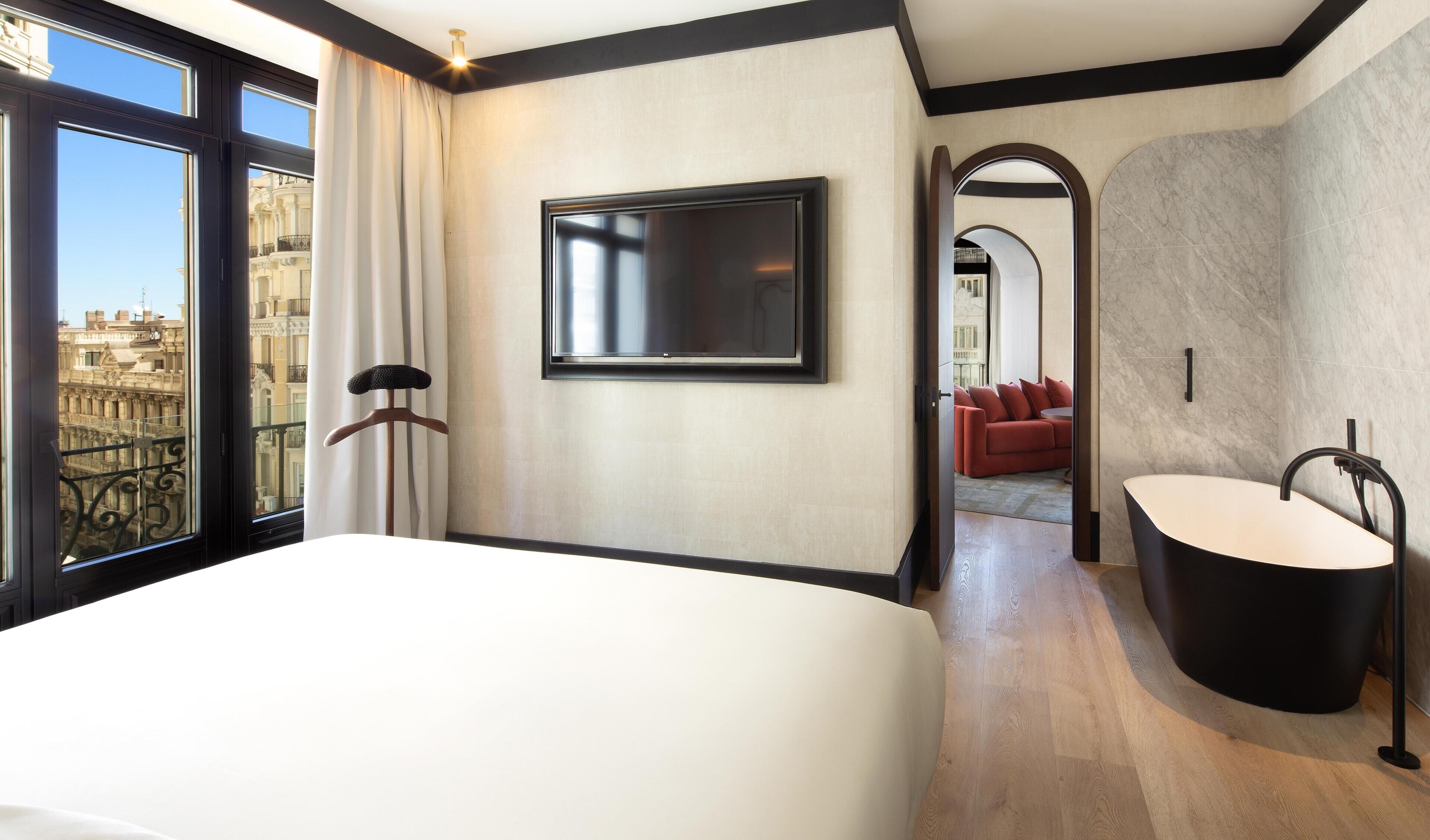 A hotel room with a large bed, a TV, a bathtub, and a view of the city.
