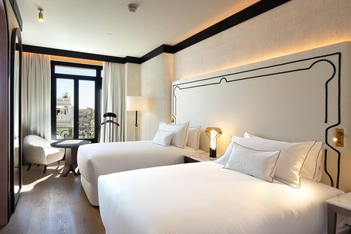A hotel room with two beds, a desk, a chair, a window with curtains, and a view of buildings.