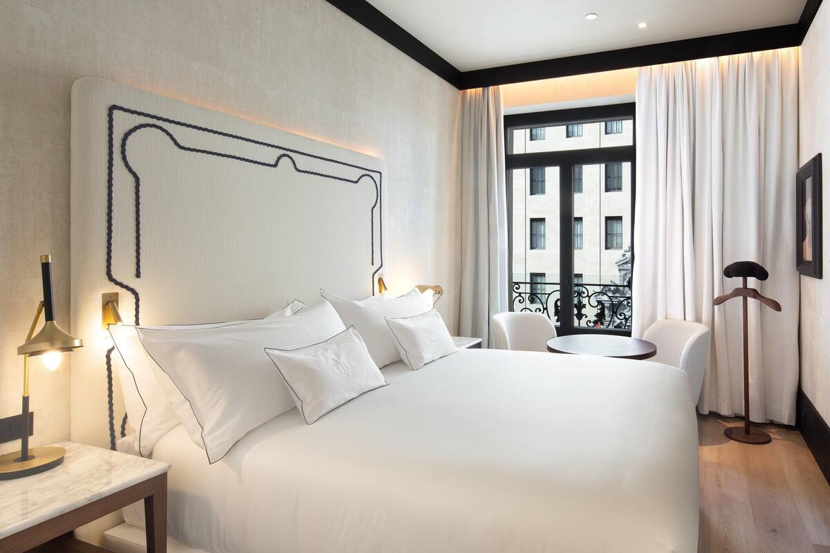 A hotel room with a large bed, bedside tables, a round table, and a view of buildings through the window.