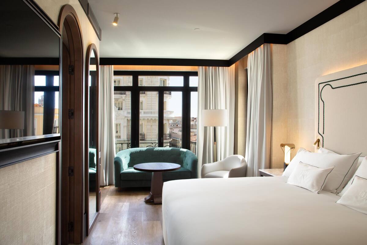 A hotel room with a bed, a round table, a green armchair, and a view of buildings through large windows.