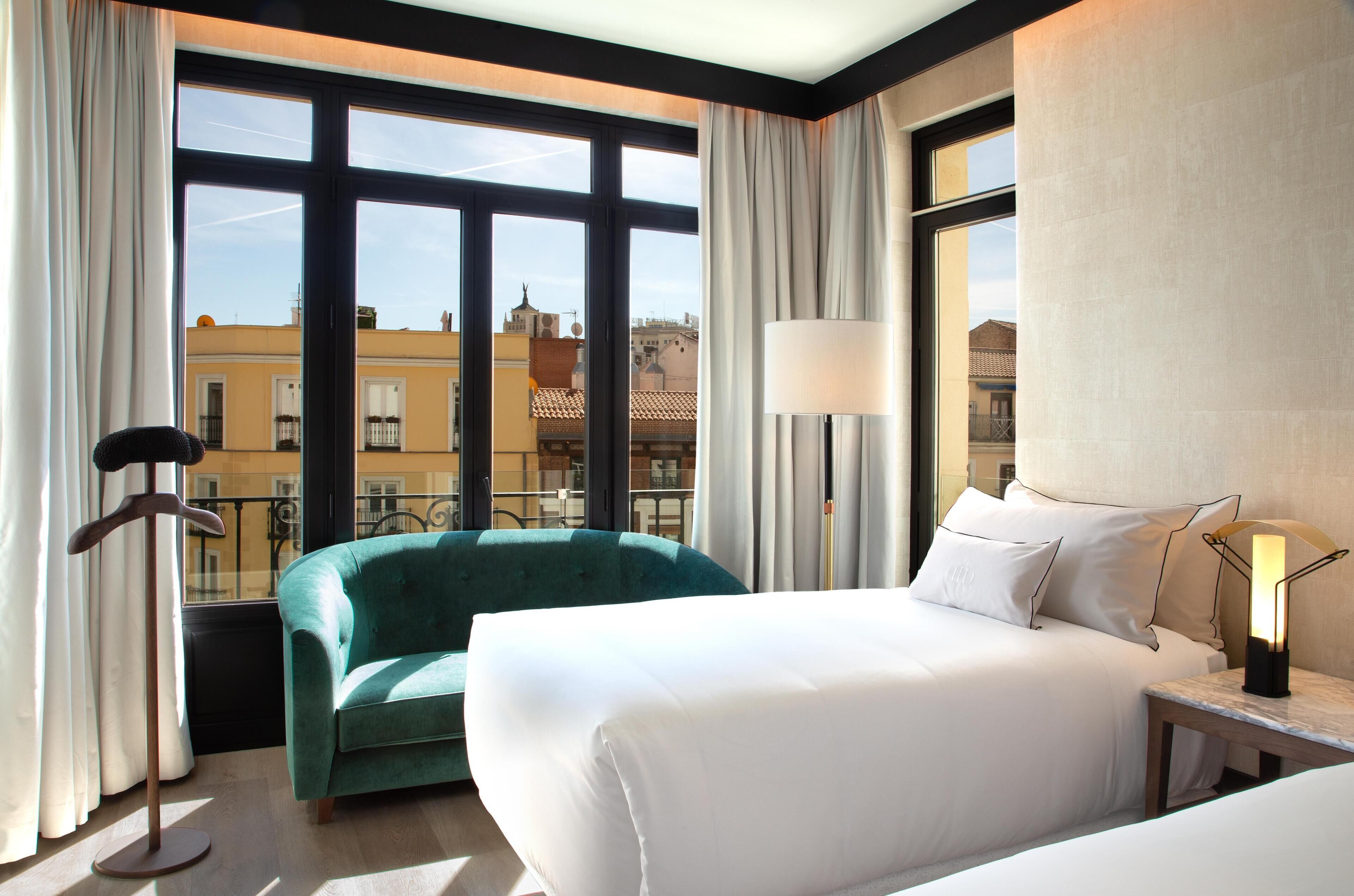 Ein Hotelzimmer mit einem Bett, einem grünen Sessel, einer Stehlampe und Blick auf Gebäude durch große Fenster.
