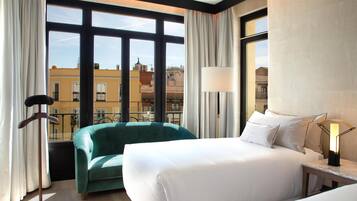 Ein Hotelzimmer mit einem Bett, einem grünen Sessel, einer Stehlampe und Blick auf Gebäude durch große Fenster.