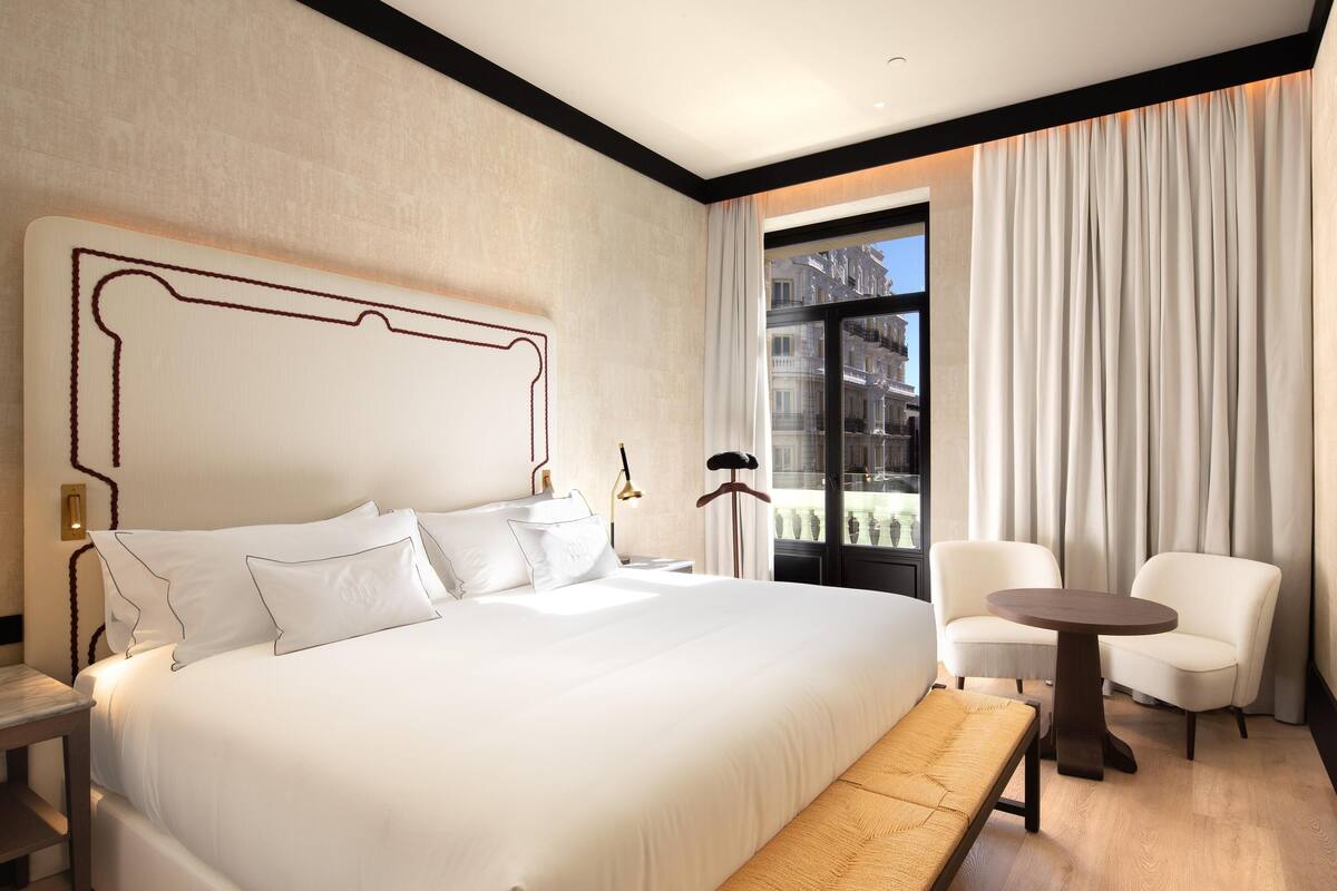 A modern hotel room with a large bed, a bedside table, a lamp, a chair, a small table, and a view of the city through a window.
