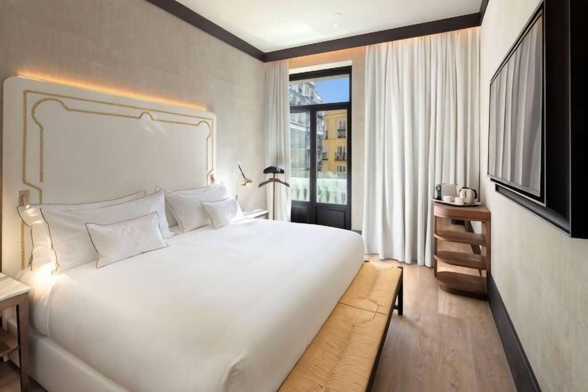 A hotel room with a large bed, a bedside table, a nightstand, a window with curtains, and a view of buildings outside.
