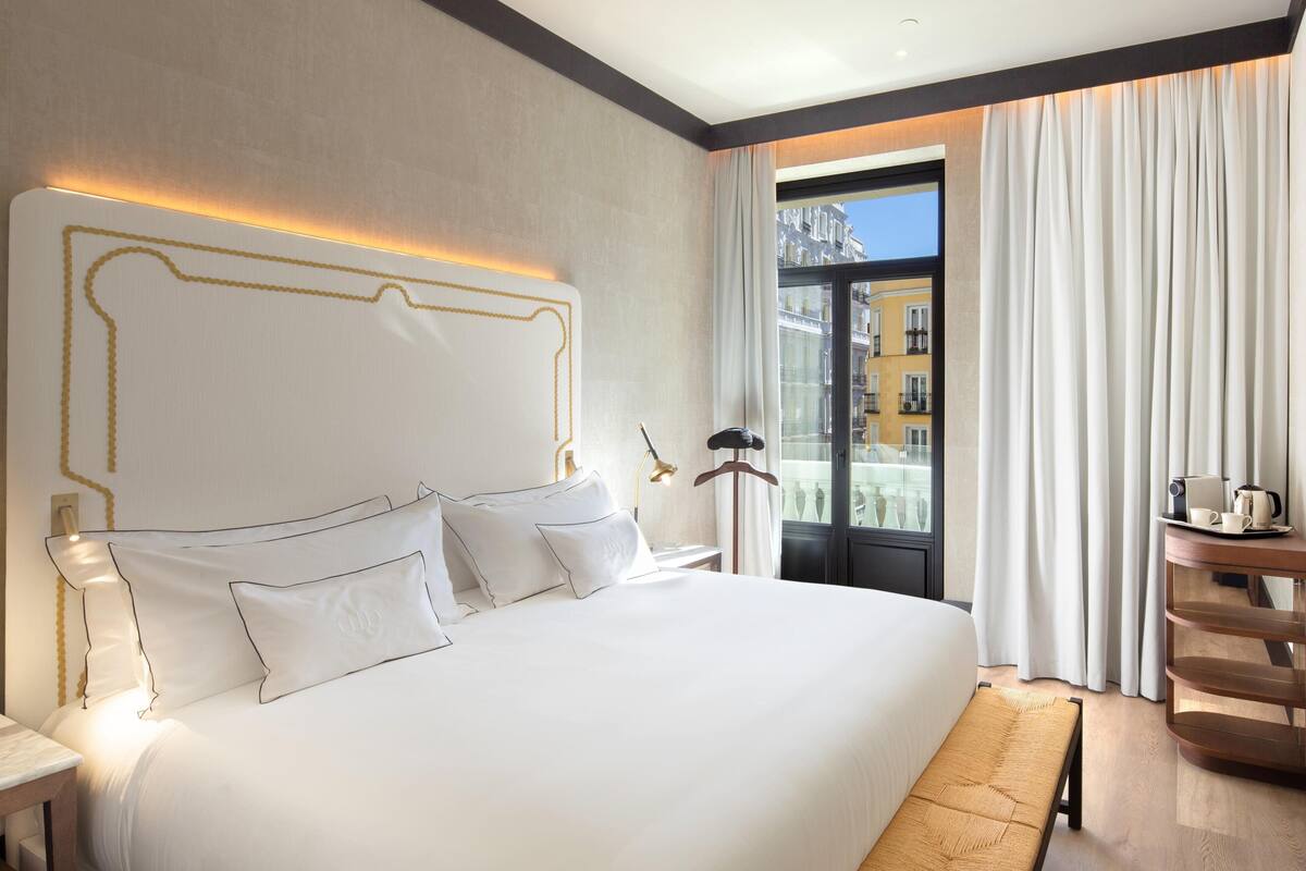 A hotel room with a large bed, a bedside table, a bench, a lamp, and a view of buildings through the window.