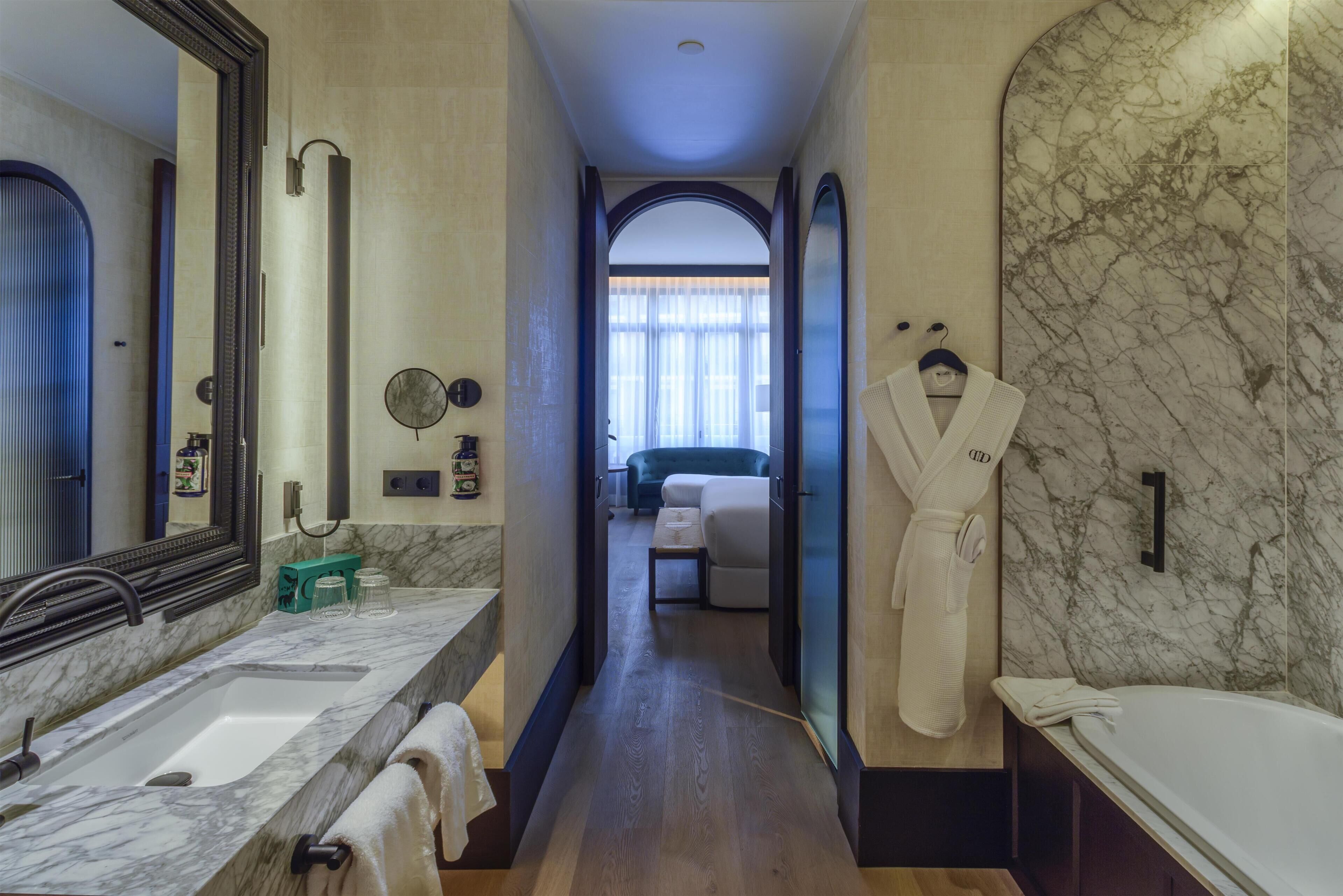 Un baño de hotel con un lavamanos de mármol, un espejo grande, una bata colgada en la pared y vistas a un dormitorio con una cama y una ventana.