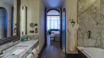 Un baño de hotel con un lavamanos de mármol, un espejo grande, una bata colgada en la pared y vistas a un dormitorio con una cama y una ventana.