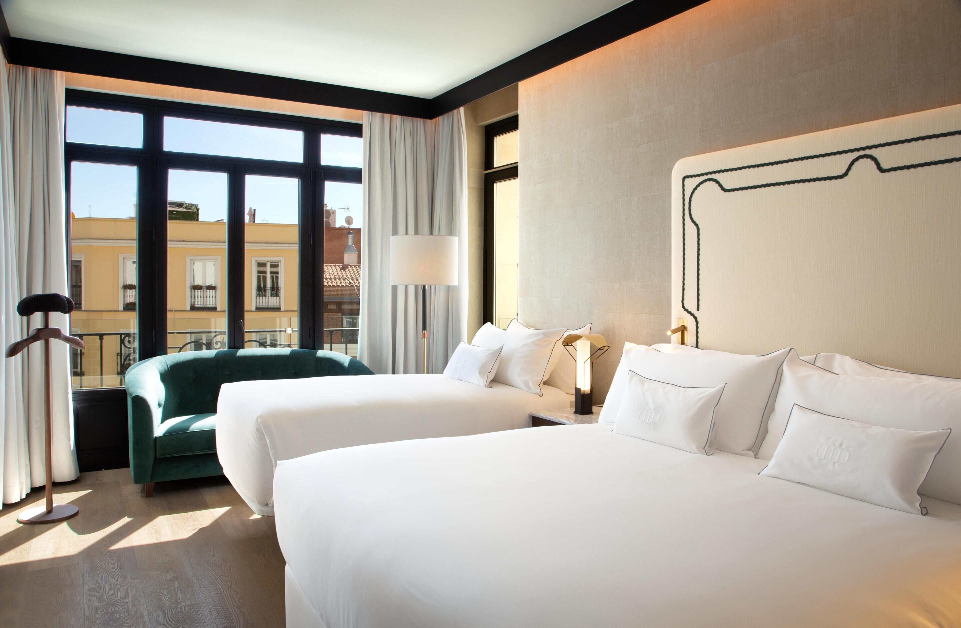 A hotel room with two beds, a green sofa, a lamp, and a view of buildings through large windows.